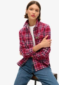 Chemise en flanelle à carreaux rouges et bleus avec des manches longues, devant à boutons, deux poches poitrine et étiquettes, portée sur un T-shirt blanc et un jean.