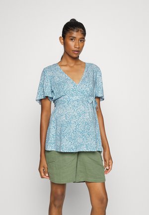 Top envolvente azul e branco com padrão, mangas curtas, decote em V e detalhe de cintura amarrada, combinado com calções verdes.
