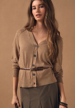 Kvinde iført en brun cardigan med knapper og bælte, plisseret mørk nederdel og armbånd, står mod en beige baggrund.