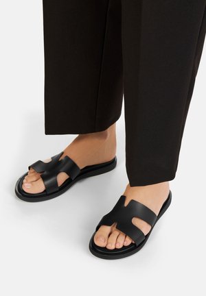 Sandali sabot neri con design traforato, realizzati in pelle liscia. Presentano una suola piatta e componenti hardware minimal per un aspetto elegante.