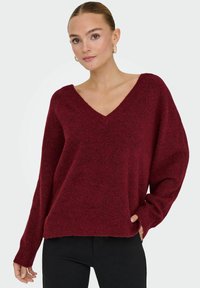 Pull en v color bordeaux avec des manches longues, texture douce, coupe ample et ourlet côtelé. Associé à un pantalon noir ajusté.