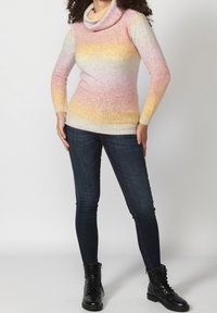 Cowl neck sweater in gradient pastel colors, featuring pink, yellow, and blue hues, with ribbed cuffs. Worn with dark fitted jeans and black boots.