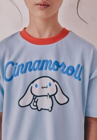 T-shirt bleu clair avec col rouge, arborant un dessin animé souriant de lapin et le texte "Cinnamoroll" en bleu, texture douce en coton.