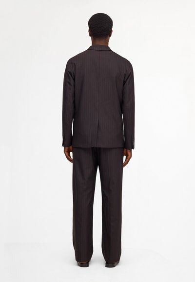 Man wearing a dark brown pinstripe suit standing facing away on a plain white background.