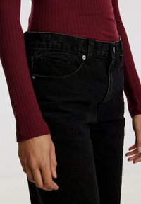 Schwarze Jeans aus Denim mit hoher Taille, ausgestattet mit Fronttaschen und einer subtilen, strukturierten Oberfläche. Kombiniert mit einem enganliegenden burgunderfarbenen Langarmoberteil.