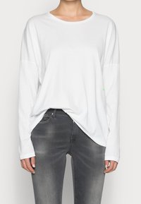 Langärmliges weißes Baumwollhemd mit rundem Ausschnitt und lockerer Passform. Kombiniert mit figurbetonten grauen Denim-Jeans, die einen subtilen Wasch-Effekt aufweisen.