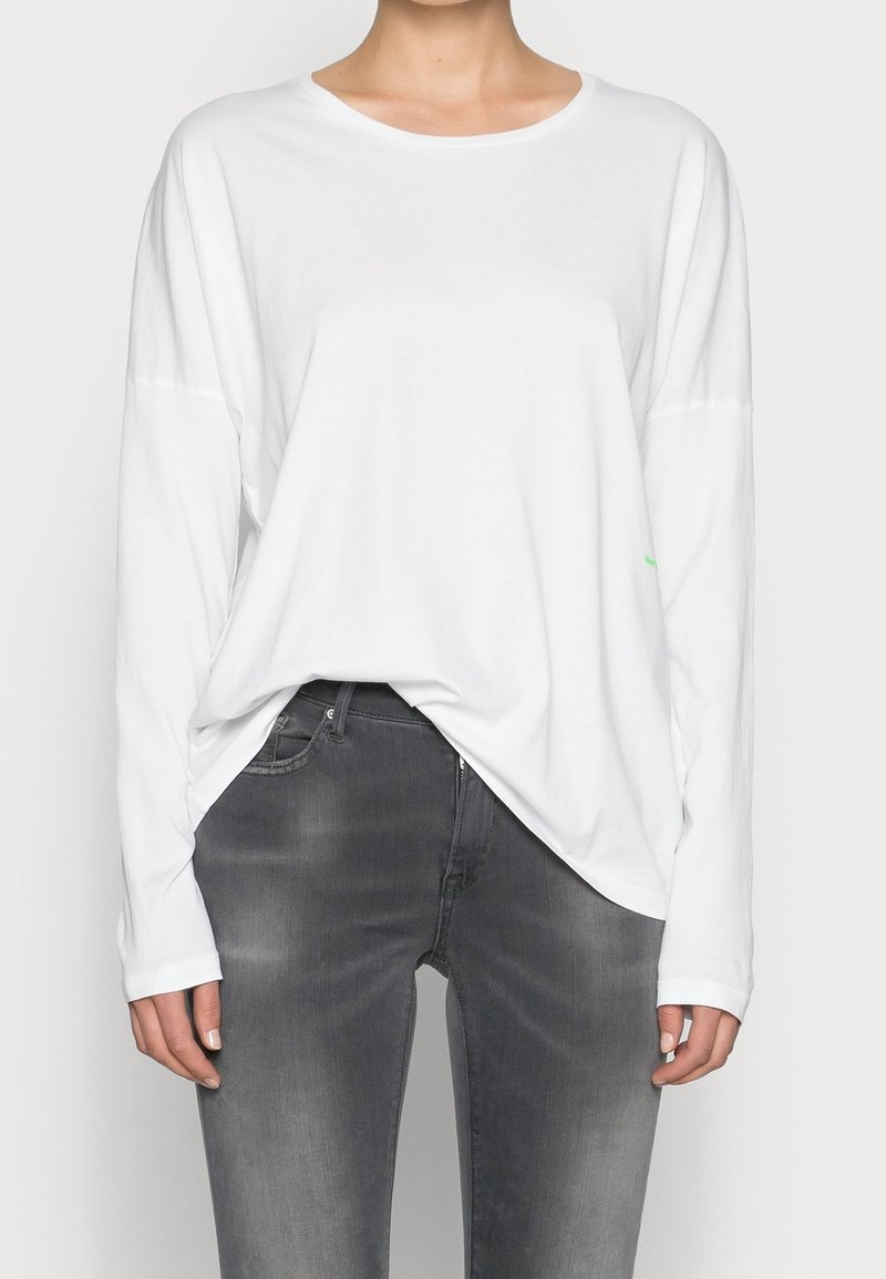 Langärmliges weißes Baumwollhemd mit rundem Ausschnitt und lockerer Passform. Kombiniert mit figurbetonten grauen Denim-Jeans, die einen subtilen Wasch-Effekt aufweisen.