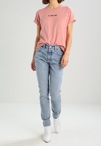 Růžové tričko s vyhrnutými rukávy a černým textem, v kombinaci se světle modrými džíny a bílými kotníkovými botami, stojící na světlejším pozadí.