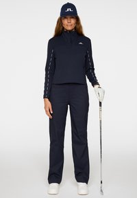 Abbigliamento da golf navy con un top a mezza zip caratterizzato da accenti bianchi, abbinato a pantaloni coordinati e un cappellino. Tiene un bastone da golf con la mano destra.