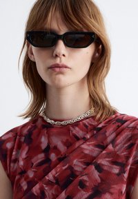 Schwarze Cat-Eye-Sonnenbrille mit dunklen Gläsern, goldenen Akzenten an den Ecken, getragen mit einem rotblumigen Oberteil und einer silbernen Kettenhalskette.