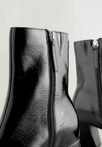 Černé kotníkové boty z lesklé lakované kůže s předním zipem a tvarovaným siluetou; mají texturovaný povrch a elegantní design.