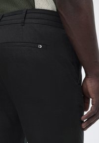 Κοντινό πλάνο ενός ατόμου που φοράει μαύρο παντελόνι με ένα μικρό λευκό λογότυπο στην πίσω τσέπη και ένα πλεκτό τοπ σε πράσινο και μπεζ χρώμα.