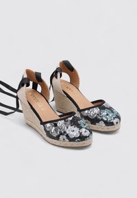 Zapatos de cuña negros con lentejuelas y diseño floral, con suela intermedia de yute y lazos de cintas negras. Parte trasera y laterales abiertos para un aspecto aireado.