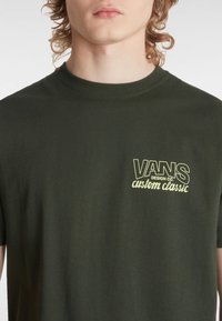 Mørkegrøn bomulds t-shirt med rund hals, der har et gult silketrykt logo, der står "VANS DESIGN CO. custom classic" på brystet.
