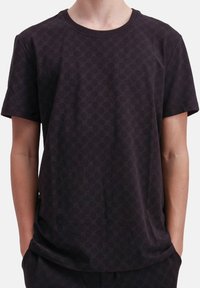 Een persoon in een donker kortarmig shirt met een subtiel cirkelpatroon, met de handen gedeeltelijk in de zakken, tegen een effen achtergrond.