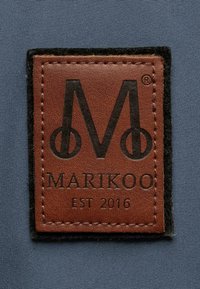 Brown leather label with black text and logo, stitched onto gray fabric. Details include the brand name "MARIKOO" and "EST 2016."