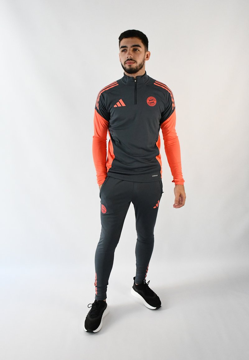 Chándal deportivo en gris oscuro con toques en naranja, que presenta un cuello con cremallera, mangas largas, pantalones ajustados y detalles de logo. Zapatillas deportivas negras.