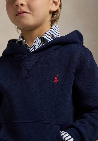 Enfant portant un sweat à capuche bleu marine avec un logo rouge brodé sur une chemise à rayures bleues et blanches avec col et petits motifs d’ours.