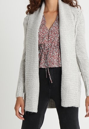 Personne portant un cardigan ouvert en maille gris clair sur un chemisier rouge et blanc à motifs avec un pantalon noir.