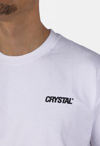 Bílý bavlněný tričko s černým potiskem loga, které zní "CRYSTAL." Kulatý výstřih se krátkými rukávy, hladká textura.