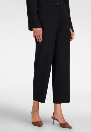 Vrouw draagt zwarte, brede cropped broek en bruine slingback hoge hakken met puntige neus, staand op een lichte achtergrond.