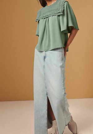 Kvinde iført en salviegrøn bluse med en bred strikket krave og en lyseblå denim maxi nederdel med en frontslids samt hvide ankelstøvler med frynser.