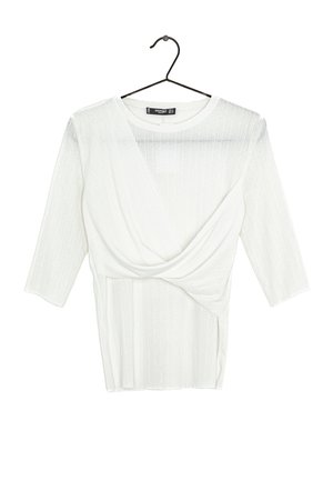 Valkoinen pitkähihainen ribbineulottu toppi, jossa on röyhelömäinen ristiin-kietoutuva yksityiskohta edessä, roikkuu mustassa vaatehenkarissa valkoista taustaa vasten.