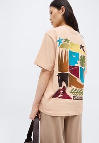 Γυναίκα με μπεζ μπλουζάκι που έχει πολύχρωμα αφηρημένα σχήματα, αστέρια και φύλλα στην πίσω πλευρά, κρατώντας τσάντα με σκούρο καφέ υφή και λουρί.