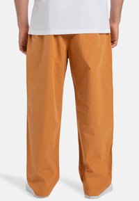 Loose-fitting, orange cotton pants with a smooth texture and subtle stitching details. Tapered towards the ankle, worn with white shoes.