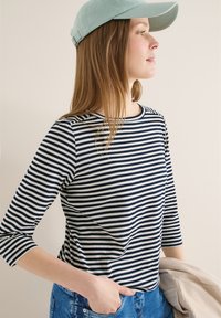 Femme portant une casquette bleu clair et une chemise rayée bleu marine, main dans la poche de son jean, tenant une veste beige sur un fond uni.