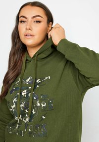 Grön bomullshuvtröja med dragsko, försedd med en grafisk design i vitt, blått och kamouflage på framsidan.