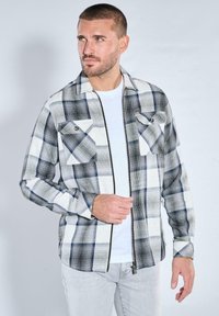 Schwarz-weiß karierte Reißverschluss-Hemdjacke mit zwei Fronttaschen und grauen Ärmeln, getragen über einem weißen T-Shirt, vor einem hellen Hintergrund.