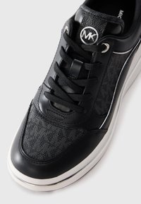Nahaufnahme eines schwarzen Michael Kors Sneakers mit strukturiertem Stoff, Lederelementen, schwarzen Schnürsenkeln und weißer Sohle vor einem hellen Hintergrund.