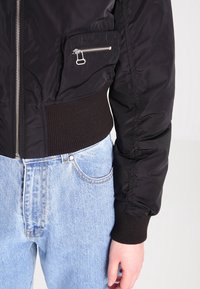 Svart bomberjacka med silvrig dragkedja och dragkedjeficka, bärs med ljusblå högmidjade jeans som visar midjan och höger arm.