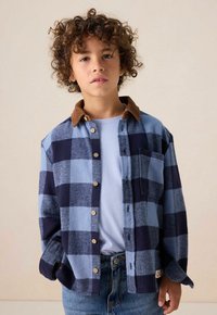 Flanellen shirt met blauwe en marinekleurige ruiten, knoopsluiting aan de voorkant, borstzak en een bruine corduroy kraag, gedragen over een lichtblauw T-shirt.