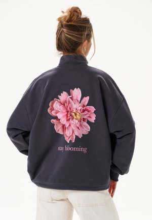 Femme avec les cheveux en chignon portant une veste sombre montrant une grande fleur rose et le texte « stay blooming » au dos, associée à un pantalon clair.