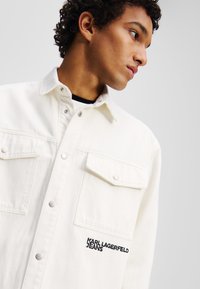 Bijela traper košulja s dva džepa na prsima, dugmad na preklapanje i crni vez "KARL LAGERFELD JEANS" na lijevom džepu. Glatka tekstura.