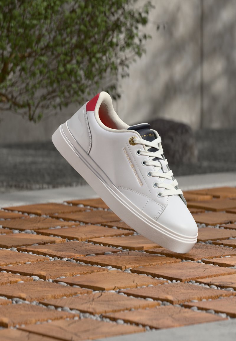Baskets blanches Tommy Hilfiger avec languette rouge au talon, œillet doré et lacets, flottant au-dessus d'une surface extérieure en carreaux de bois et gravier.