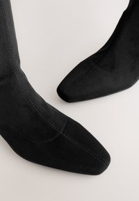 Bottes cheville en daim noir avec bouts pointus, côtés incurvés et détails de couture subtils. Texture lisse, design minimaliste.