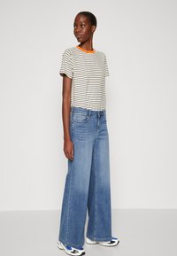 Camiseta de rayas con cuello naranja, combinada con unos vaqueros de pierna ancha azul. El modelo lleva zapatillas blancas y negras con detalles en azul.