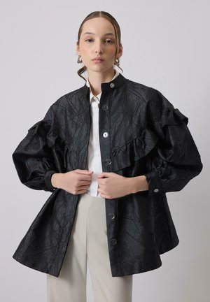 Młoda kobieta ubrana w czarną, teksturowaną kurtkę z falbanami i wysokim kołnierzem, nałożoną na białą koszulę i beżowe spodnie, stojąca na jednolitym tle.