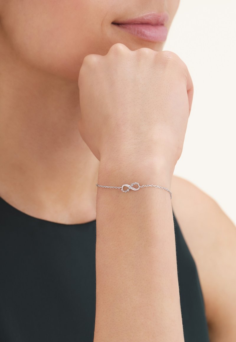 Femme portant un délicat bracelet en chaîne argentée avec deux charms en forme de cœur reliés autour de son poignet, reposant son poing contre son menton.