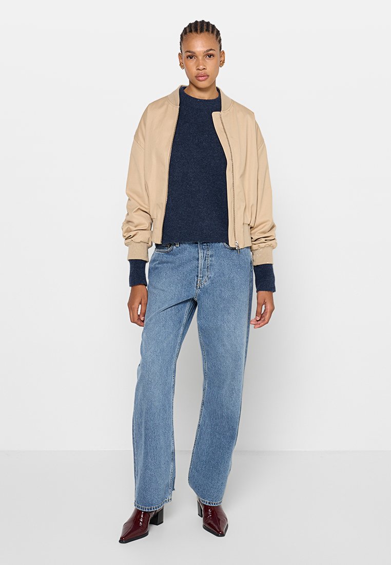 Beige Bomberjacke über einem dunklen Pullover, kombiniert mit hellblauen, weiten Jeans und burgunderfarbenen Stiefeletten. Einfaches Design mit lässiger Passform.