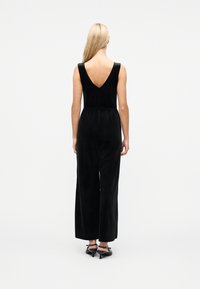 Γυναίκα με μακριά ξανθά μαλλιά που φοράει μαύρη αμάνικη ολόσωμη φόρμα με βαθύ V στην πλάτη και μαύρα πέδιλα με τακούνι και λουράκι, στέκεται μπροστά σε λευκό φόντο.