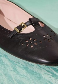 Chaussure Mary Jane en cuir noir avec motif floral découpé et boucle dorée sur une surface turquoise texturée.