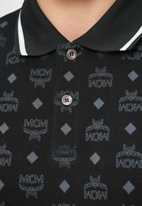 Polo negro con tela texturizada con patrón repetido de logotipo MCM y acentos de diamante gris, acabado con un cuello de contraste.