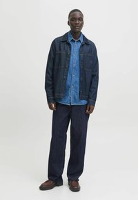 Chaqueta de mezclilla oscura con bolsillos frontales sobre una camisa de mezclilla más clara, combinada con jeans anchos oscuros y zapatos marrones con cordones.