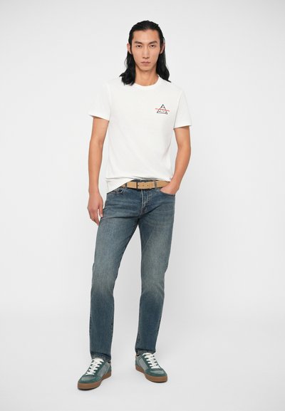 Valkoinen puuvillanen T-paita, jossa on pieni vuorilogot, yhdistettynä sinisiin farkkuihin ja vihreisiin lenkkareihin, joissa on valkoiset nauhat ja ruskeat pohjat.