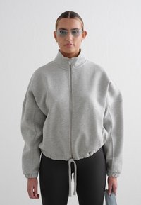 Grå hettegenser med glidelås, høy krage, elastisk snor nederst og ribbede mansjetter, kombinert med smale svarte leggings og tonede briller.