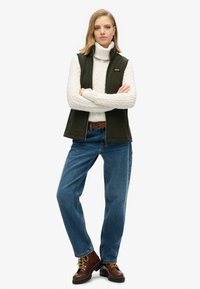 Zielona polarowa kamizelka noszona na białym golfie o splocie w warkocze, zestawiona z niebieskimi dżinsami i brązowymi butami na sznurówki. Prosta klamra, luźny krój.